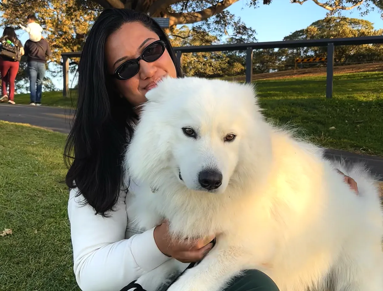 Woman with long dark hair and sunglasses hugs a large, fluffy white dog—her recent adoption—while sitting on grass in a park, giving the pup a new lease on life. Trees and people fill the background.