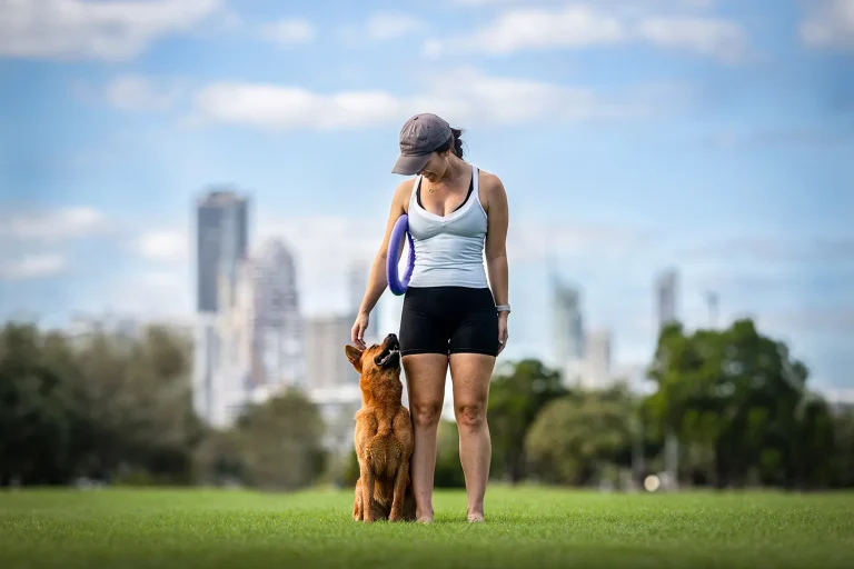 A woman, April Butler, stands on grass holding a frisbee, looking down at a Red Cattle Dog named Winnie sitting by her side. City buildings and trees are visible in the blurred background under a blue sky.